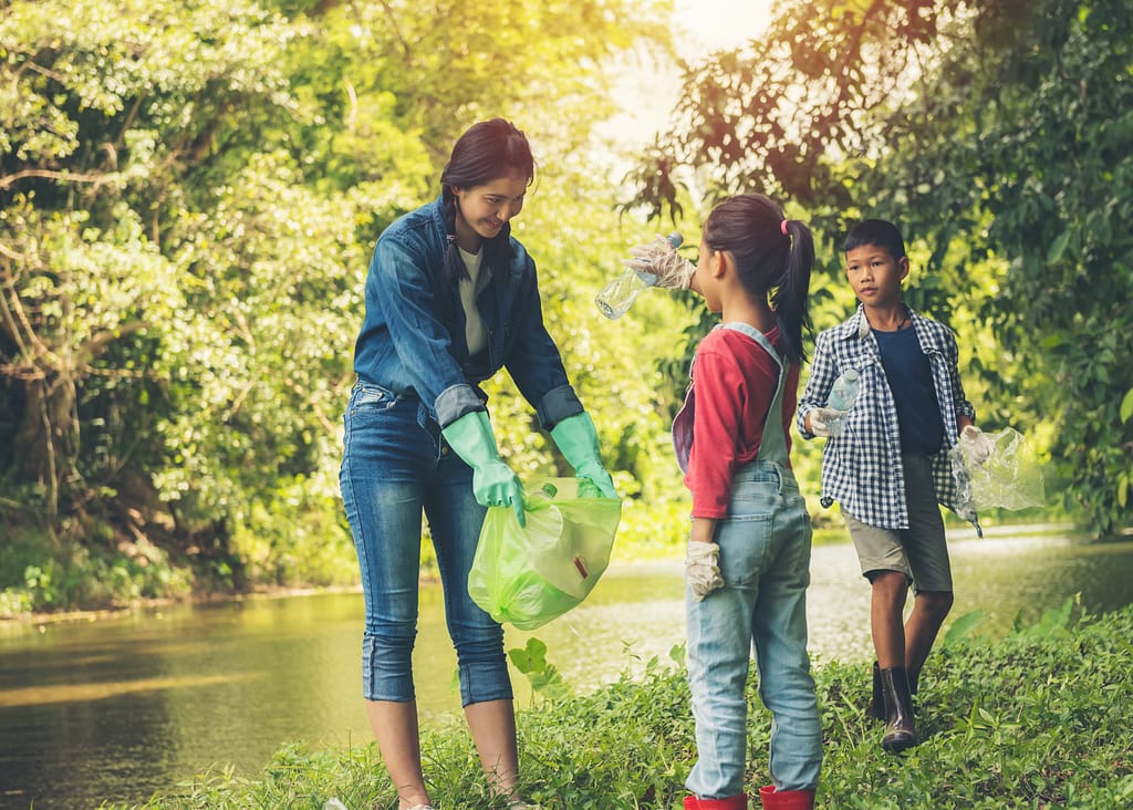 A woman with dark hair holding open a trash bag while two children pick up garbage in a park